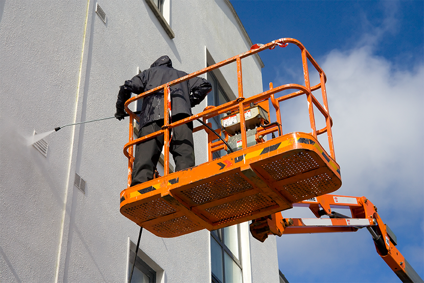 Worker in lift pressure washing building in protective gear.